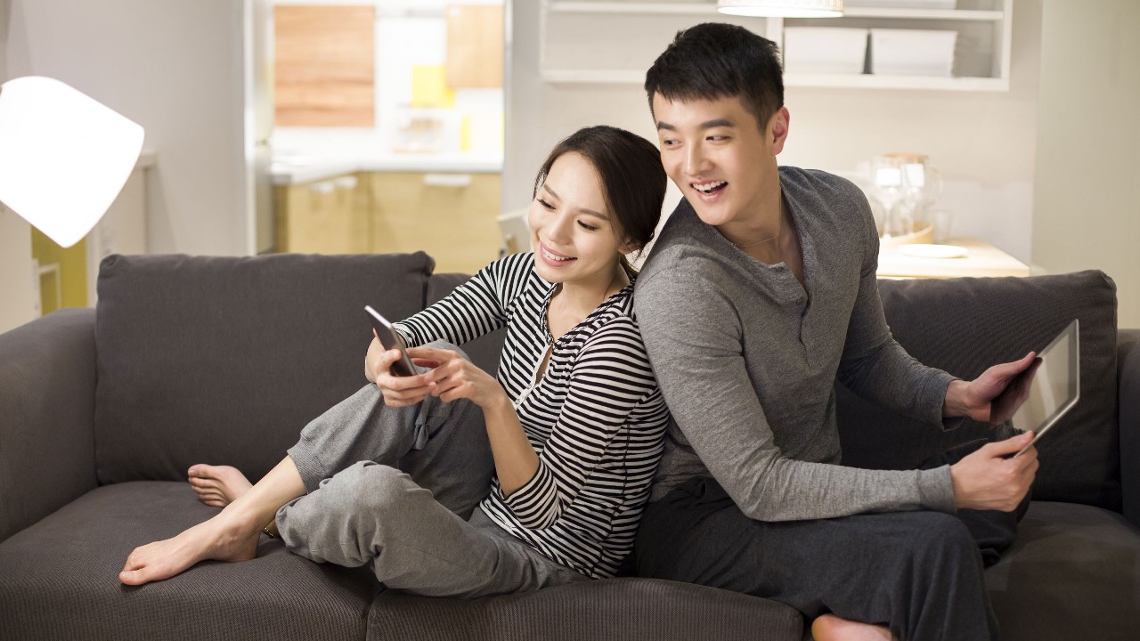 Đôi vợ chồng đang ngồi trên ghế sofa sử dụng thiết bị điện tử