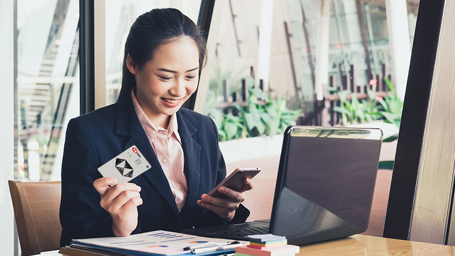 A woman is sitting on a chair, holding a credit card on her hand and looking at her laptop