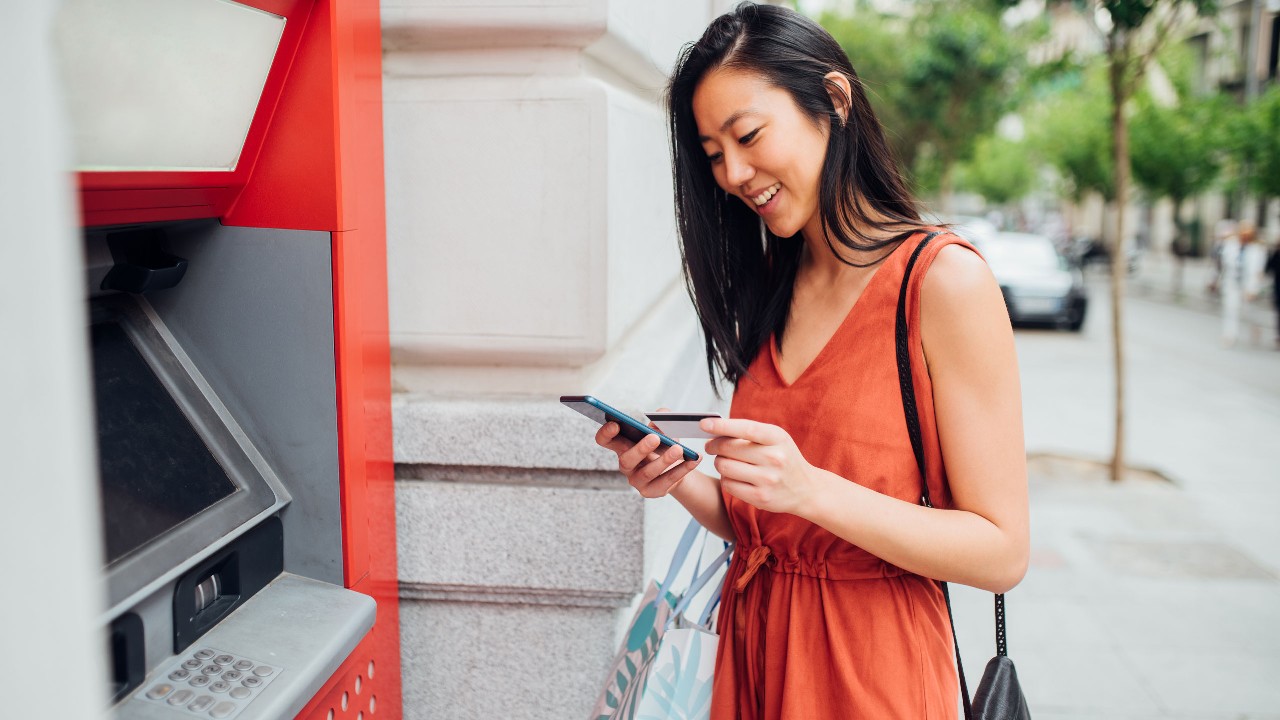 A woman is looking at her mobile phone and bank card in front of an ATM machine, image used for hsbc cash advance page