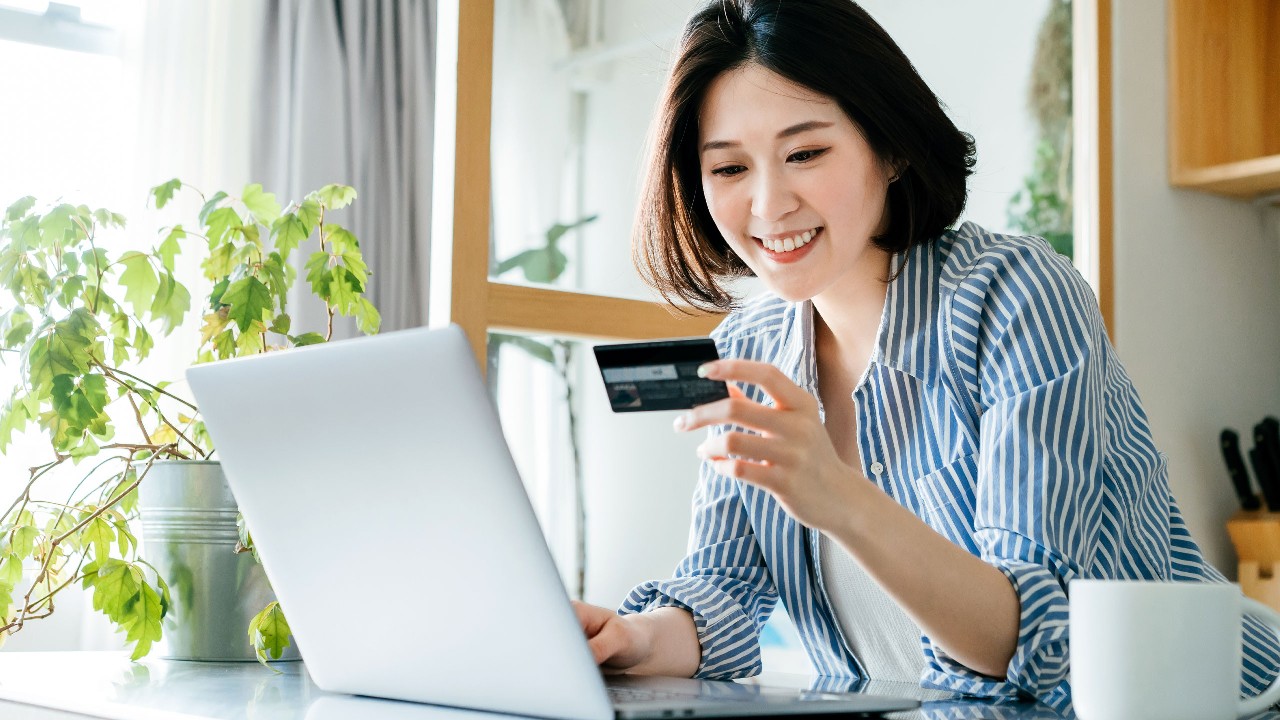 A woman is looking at her credit card and typing on her laptop