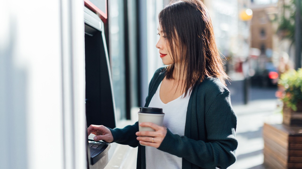 A woman is using the ATM machine while holding a cup of coffee