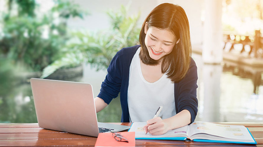 A woman writing in notebook with a laptop aside; image used for how to avoid credit card fees article page.