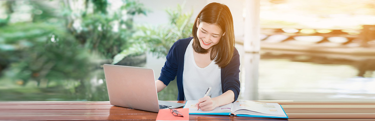 A woman writing in notebook with a laptop aside; image used for how to avoid credit card fees article page.